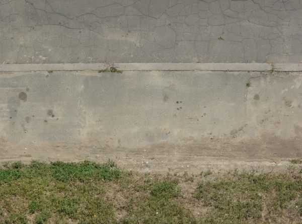 Road edge texture, with a concrete surface covered in fine dust in between a cracked dark road and an area of weeds and dirt.